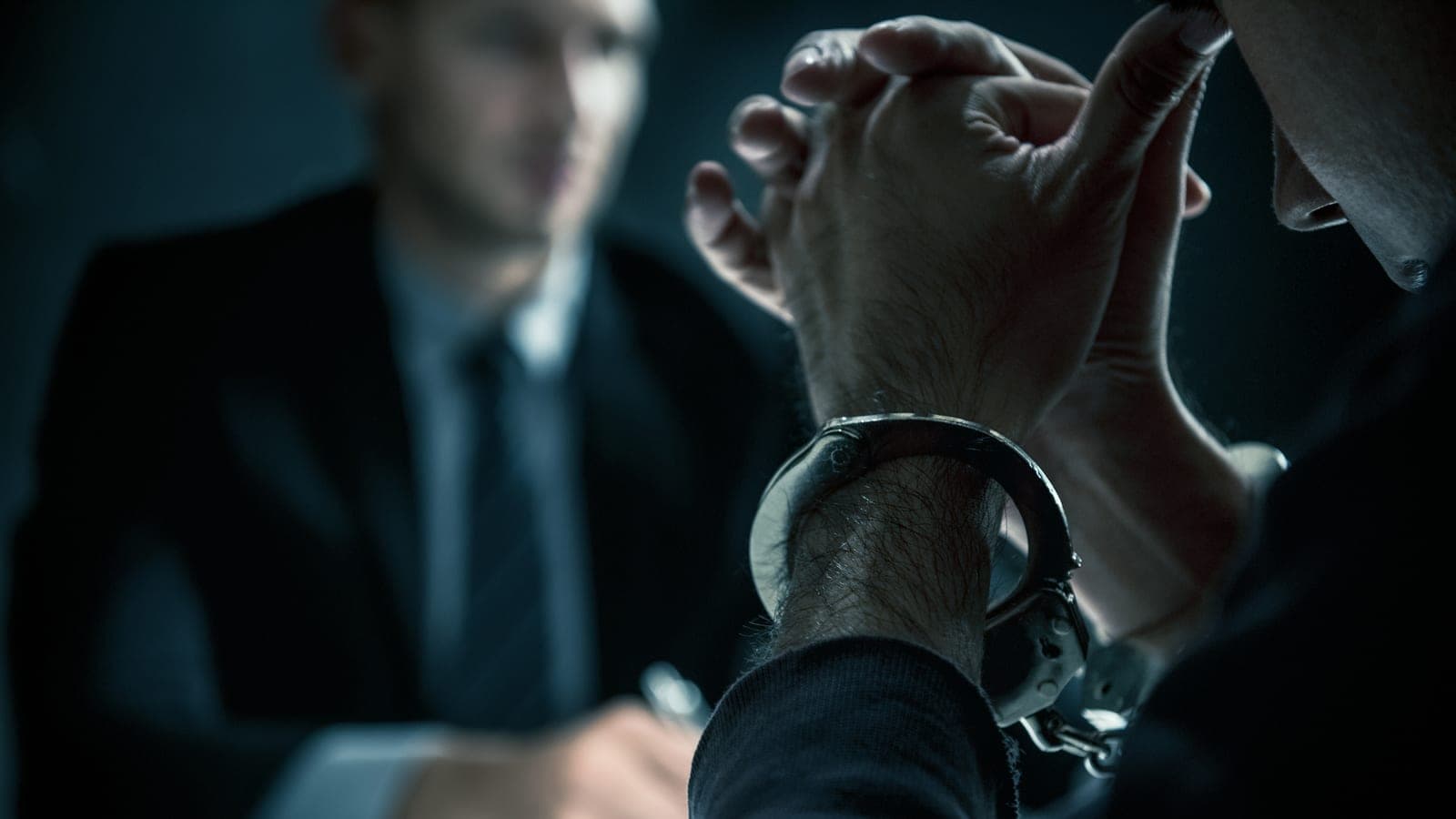 A handcuffed person seated across a table from a lawyer in a dim interview room