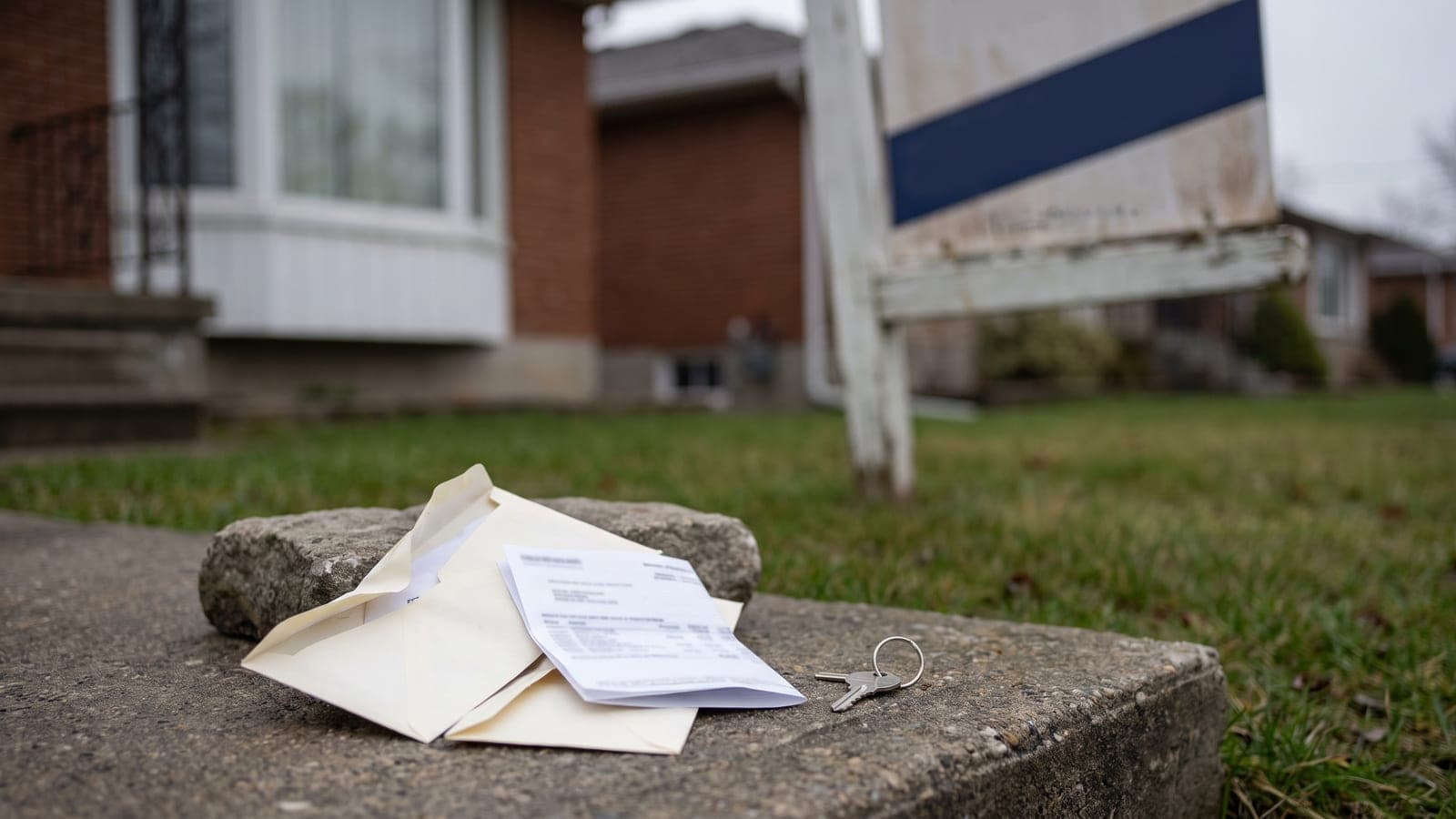 A sold sign on the lawn of an Ontario house with a stack of opened envelopes and a bank statement on the porch step